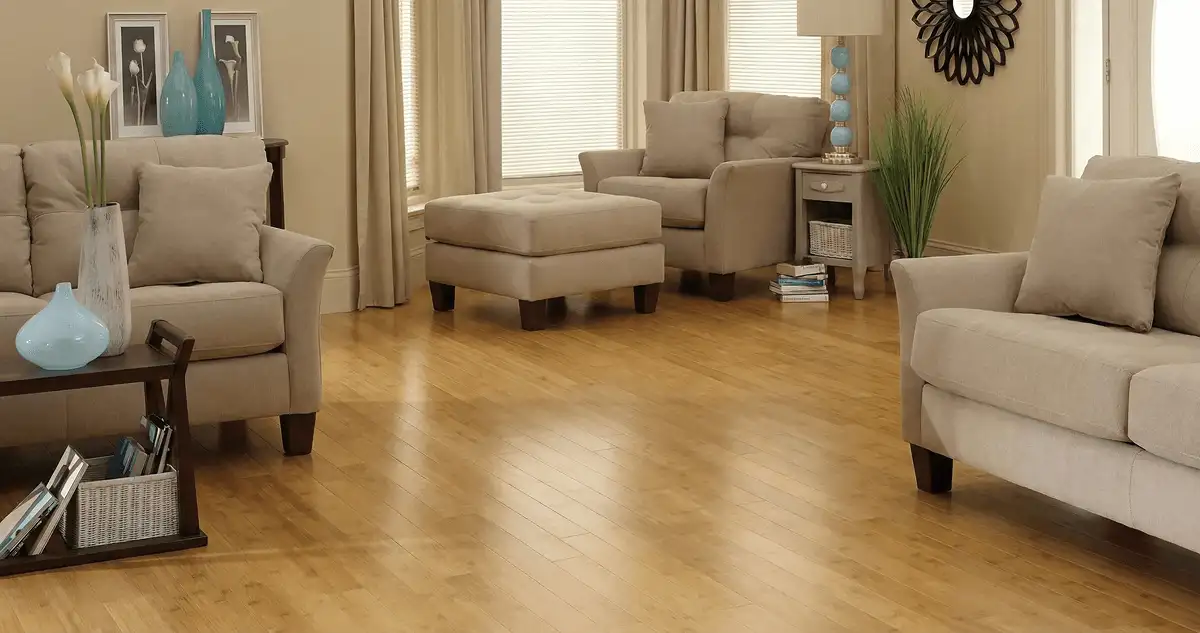 A living room with professionally installed bamboo flooring, beige sofas, a matching ottoman, a side table with a blue lamp, vases, framed pictures, and large windows with blinds and curtains in Detroit, MI